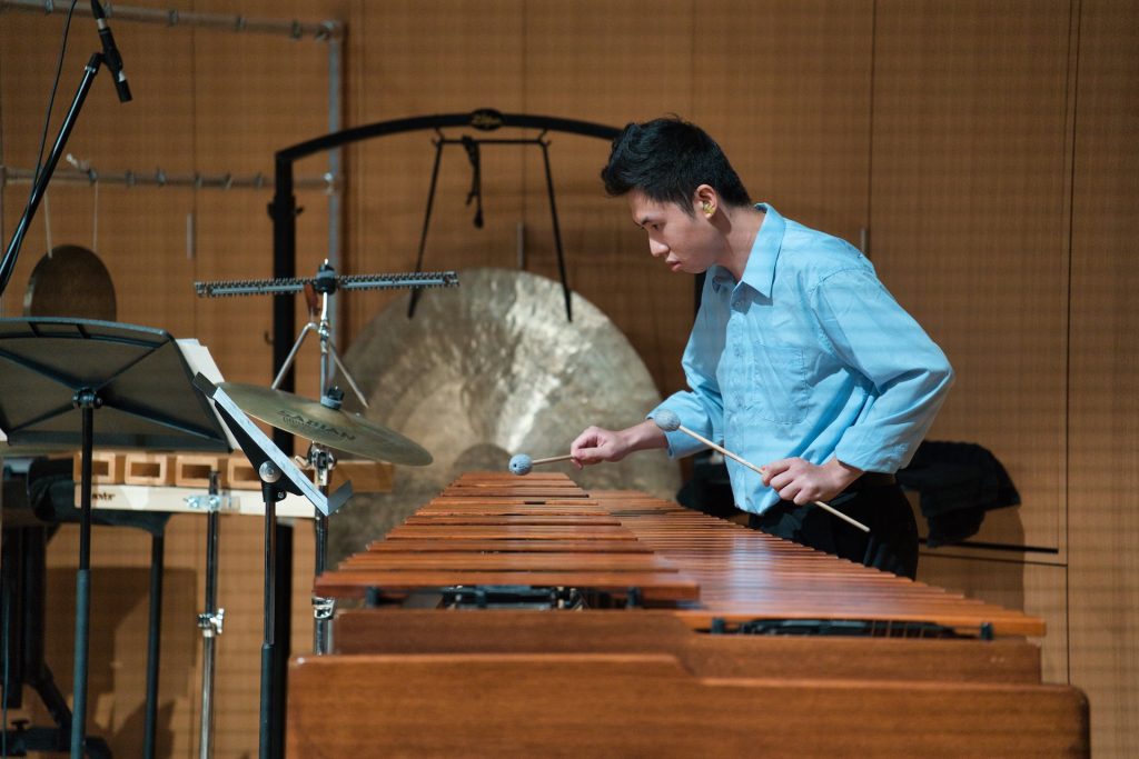 A musician in a blue shirt plays a marimba with intensity, surrounded by percussion instruments, including a gong, in a formal setting.